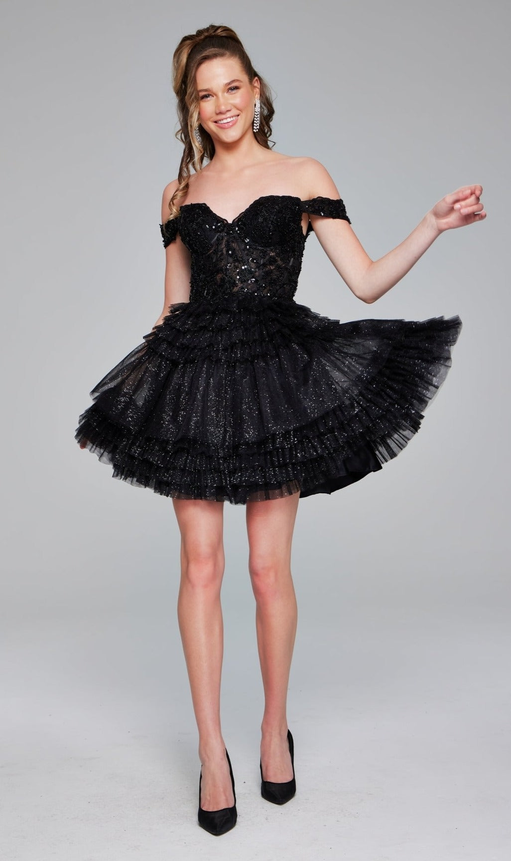 A woman smiles against a plain background in the Short Homecoming Dress 40612 by Jovani, featuring an off-the-shoulder neckline, ruffled skirt, and black heels.